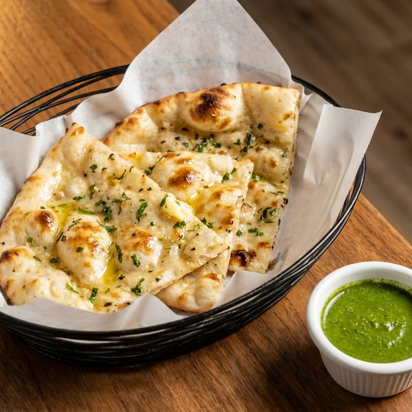 Garlic naan bread in a basket with a side of green chutney on a wooden table.