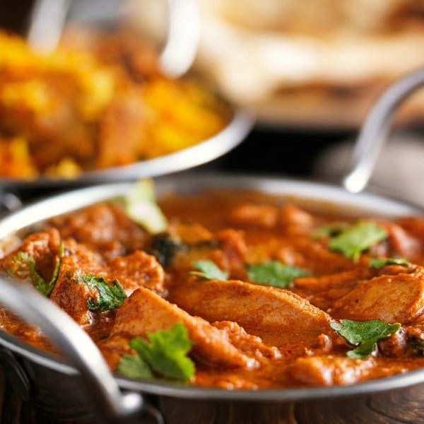 Chicken tikka masala in a metal bowl, with rice and naan bread blurred in the background.