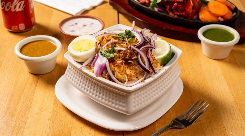 Biryani with egg, red onions, and lime, served with sauces and a soda on a wooden table.